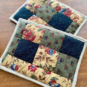Puede incluir: Dos salvamanteles acolchados con un diseño de patchwork. Cada salvamanteles presenta cuadrados de telas florales y lisas en tonos beige, verde y azul marino. Los bordes están acabados con un ribete beige claro.