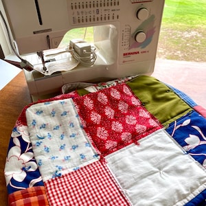 May include: A white Bernina sewing machine, displaying various stitch settings, sits above a patchwork quilt. The quilt is composed of squares in red, blue, green, and white, with floral and gingham designs. The machine is connected to a power supply and ready for sewing.