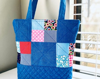 May include: A blue denim tote bag with a patchwork design. The patchwork includes various fabrics with floral, polka dot, and plaid patterns in pink, red, and blue.