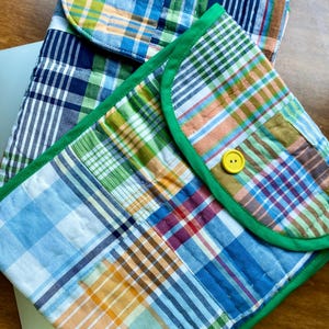 Puede incluir: Dos fundas acolchadas para portátiles con estampados de cuadros en tonos azules, verdes, naranjas y blancos. Una funda tiene un borde verde y un cierre de botón amarillo. Las fundas están sobre una superficie de madera.