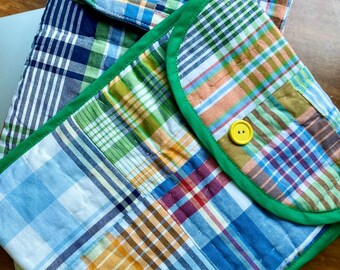 May include: Two quilted laptop cases with plaid patterns in shades of blue, green, orange, and white. One case has a green border and a yellow button closure. The cases are on a wooden surface.