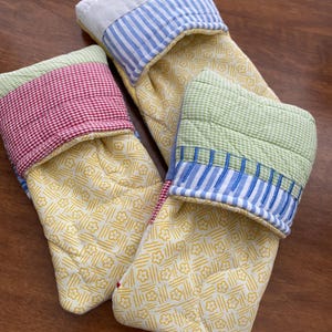 Könnte beinhalten: Drei gesteppte Ofenhandschuhe. Die Handschuhe haben ein gelbes Blumenmuster auf dem Hauptteil. Die Manschetten bestehen aus rot-weiß kariertem, blau-weiß gestreiftem und grün-weiß kariertem Stoff. Die Handschuhe liegen auf einer Holzoberfläche.