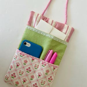 May include: A handmade fabric tote bag with a pink rope handle. The bag features a pink and white striped top, a green polka dot middle section, and a floral patterned pocket. A blue phone and pink pens are visible.
