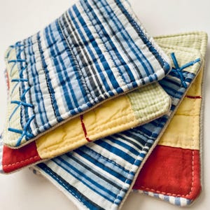 May include: Four square fabric coasters in a stack. The patchwork design incorporates blue, yellow, red, and green fabrics. Each coaster has a quilted texture and is stitched with blue thread. The coasters are approximately 10 cm x 10 cm.