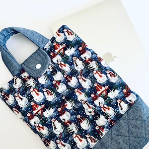 Puede incluir: Una funda azul para portátil con un estampado repetido de muñecos de nieve con bufandas rojas y sombreros de copa negros. La funda tiene un asa de mezclilla y una base de mezclilla acolchada. Un portátil plateado es parcialmente visible.