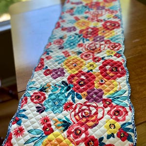 May include: Quilted floral table runner featuring a vibrant pattern of red, yellow, and turquoise flowers on a cream background. The runner has a diamond-stitched pattern and a blue patterned border. This decorative piece is perfect for adding color to a dining table.