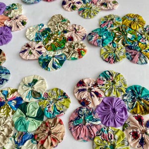 以下が含まれることがあります： 様々なサイズと模様のカラフルな布製ヨーヨーのコレクション。ヨーヨーは、ピンク、青、緑、黄色の色合いで花柄と抽象的なデザインが特徴です。これらの手作りの布の円は白い表面に配置されています。