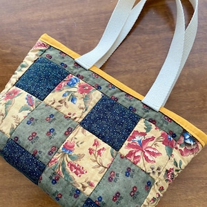 May include: A quilted tote bag with a patchwork design. The bag features a cream-colored canvas handle and a patchwork pattern of floral and solid squares in shades of blue, green, and cream. The top edge is trimmed with yellow.