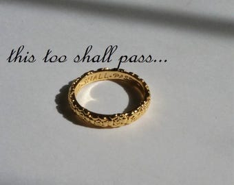 Può includere: Un anello color oro caratterizzato da un motivo floreale ripetuto lungo la fascia esterna. L'interno dell'anello è inciso con il testo "THIS TOO SHALL PASS", riportato anche in corsivo nero sopra l'anello nell'immagine.