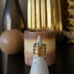 Pu&ograve; includere: Una pila di anelli color oro su un cono di cemento grigio. Gli anelli sono sottili e lucenti, che riflettono la luce. Lo sfondo include una candela sfocata e oggetti decorativi, creando un'estetica calda. Gli anelli sono un gioiello semplice ed elegante.