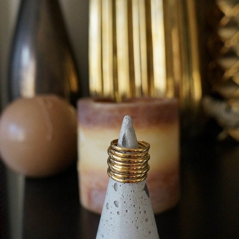 Pu&ograve; includere: Una pila di anelli color oro su un cono di cemento grigio. Gli anelli sono sottili e lucenti, che riflettono la luce. Lo sfondo include una candela sfocata e oggetti decorativi, creando un'estetica calda. Gli anelli sono un gioiello semplice ed elegante.