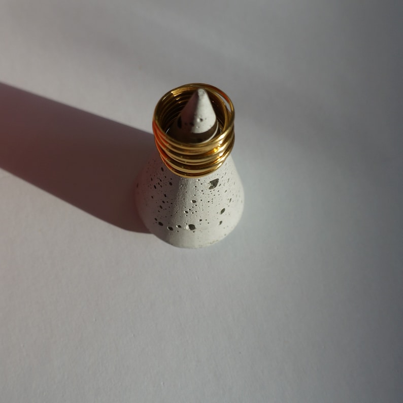 Pu&ograve; includere: Una pila di anelli color oro esposta su un piccolo portanelli in cemento macchiato. Il supporto &egrave; a forma di cono e poggia su una superficie bianca. Gli anelli sono sottili e lucenti, riflettendo la luce.
