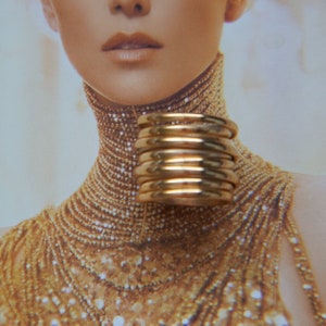 Pu&ograve; includere: Un anello largo a pi&ugrave; fasce realizzato in metallo lucido color oro. Il design presenta diversi cerchi impilati, lisci e arrotondati, che creano un aspetto continuo e stratificato sul dito quando viene indossato come pezzo unico.