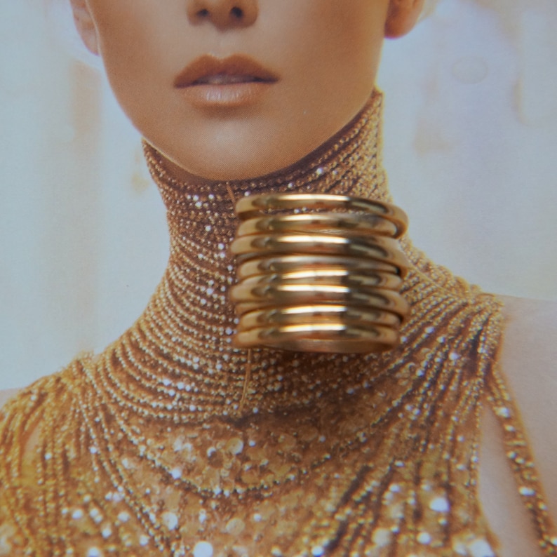 Pu&ograve; includere: Un anello largo a pi&ugrave; fasce realizzato in metallo lucido color oro. Il design presenta diversi cerchi impilati, lisci e arrotondati, che creano un aspetto continuo e stratificato sul dito quando viene indossato come pezzo unico.