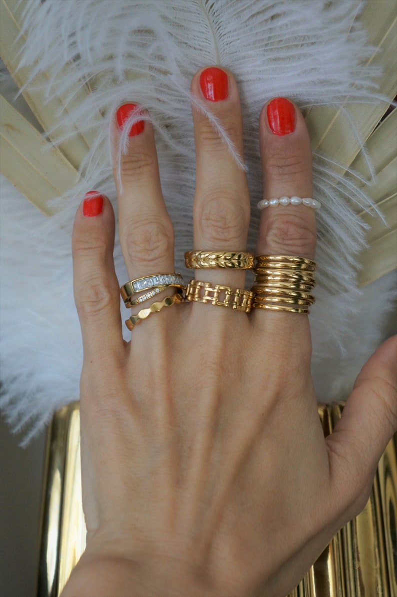Pu&ograve; includere: Una mano con diversi anelli d'oro, tra cui un anello di perle, una fascia con pietre trasparenti e pi&ugrave; anelli impilabili. Le unghie sono dipinte con smalto rosso. Lo sfondo comprende piume bianche e un vaso d'oro.