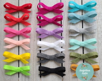 May include: A collection of colorful felt bows in various shades, including red, pink, blue, and green. The bows are arranged on a light wooden surface, showcasing their different colors and textures. The bows are a craft item.
