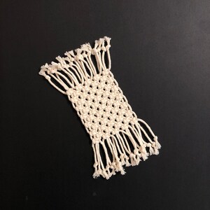 May include: A white macrame coaster with fringe on all sides. The coaster is made with a simple knot pattern.