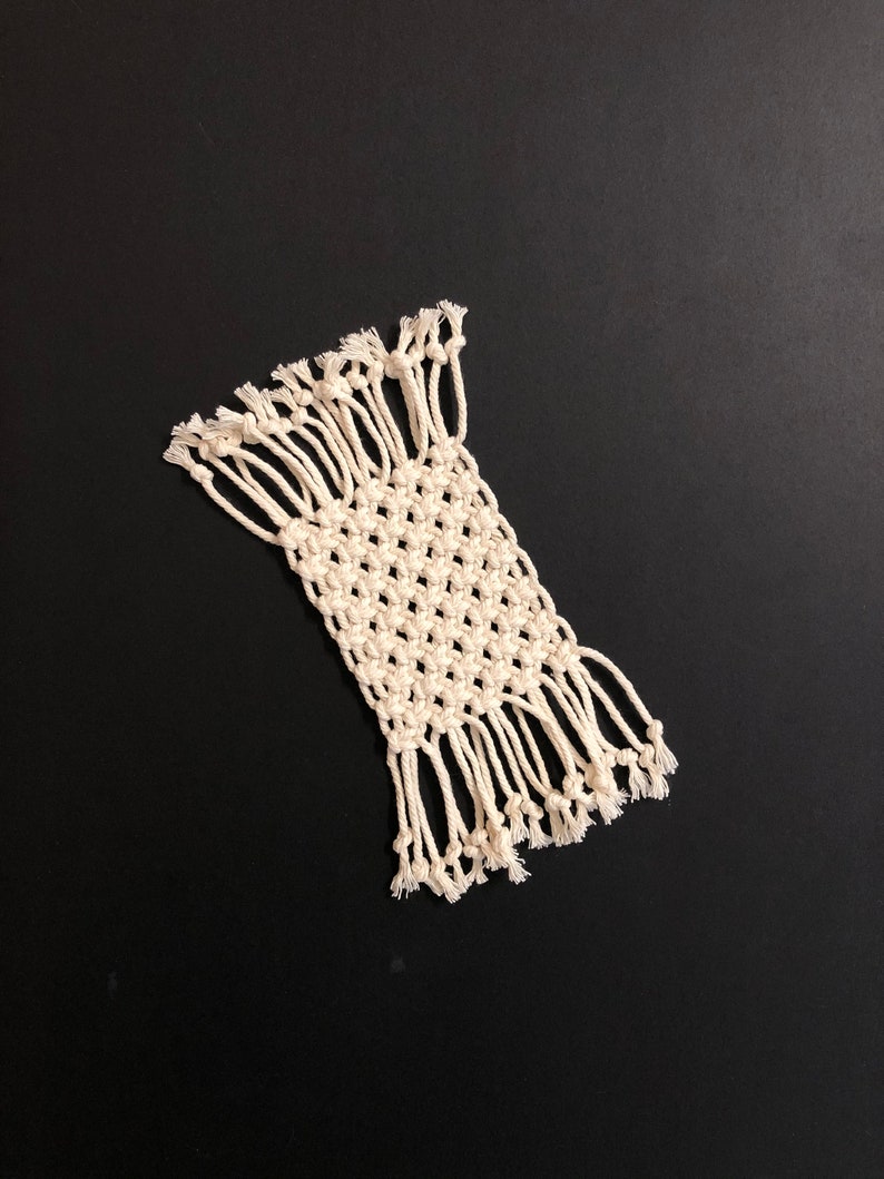 May include: A white macrame coaster with fringe on all sides. The coaster is made with a simple knot pattern.