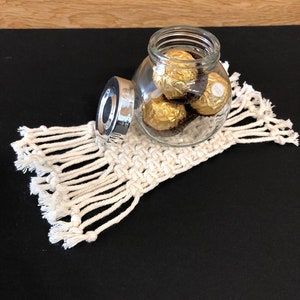 May include: A glass jar with a silver lid filled with gold-wrapped chocolate candies. The jar is sitting on a white crocheted coaster with fringe.