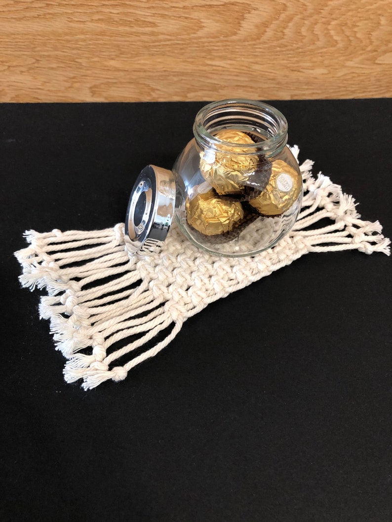 May include: A glass jar with a silver lid filled with gold-wrapped chocolate candies. The jar is sitting on a white crocheted coaster with fringe.