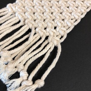 May include: A white macrame wall hanging with a fringe border. The macrame is made with a thick, natural fiber rope.