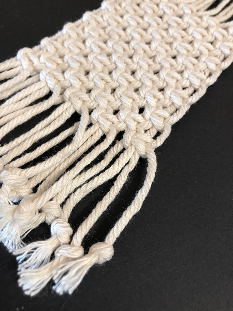 May include: A white macrame wall hanging with a fringe border. The macrame is made with a thick, natural fiber rope.