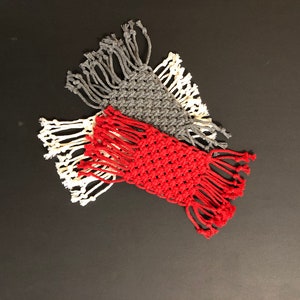 May include: Three handmade macrame coasters in red, gray, and white. The coasters are made with cotton cord and have a fringe border.