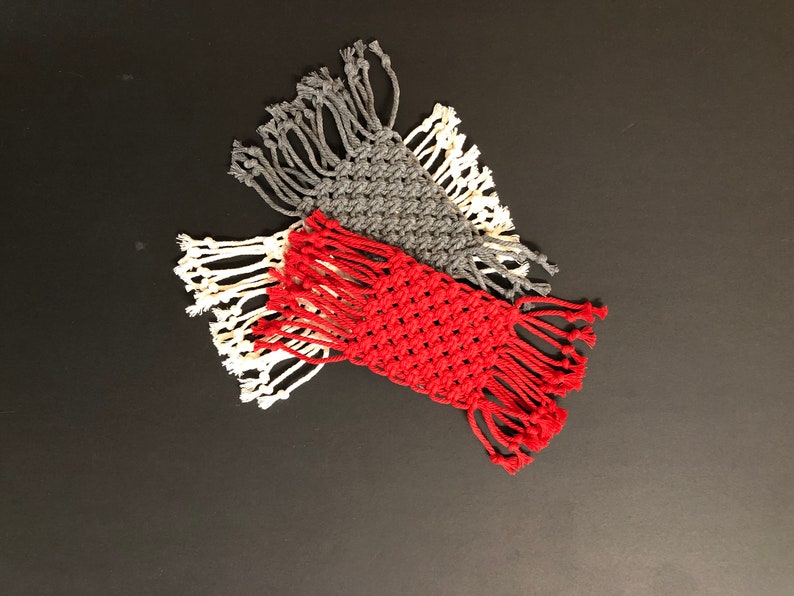 May include: Three handmade macrame coasters in red, gray, and white. The coasters are made with cotton cord and have a fringe border.