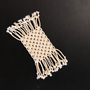 May include: A white macrame coaster with a square design and fringe edges. The coaster is made with natural cotton cord and has a simple, minimalist style.