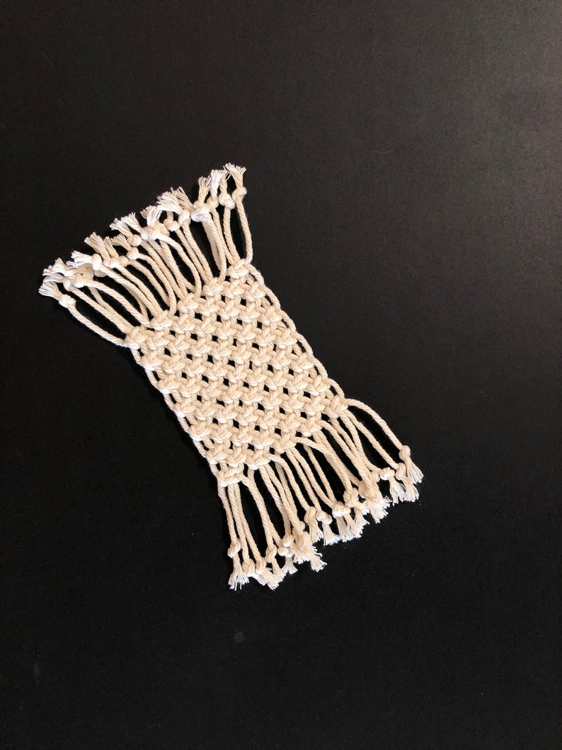 May include: A white macrame coaster with a square design and fringe edges. The coaster is made with natural cotton cord and has a simple, minimalist style.
