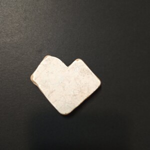 Può includere: Una piccola piastrella in ceramica a forma di cuore, bianco sporco, con una superficie screpolata. La piastrella ha una forma quadrata con un intaglio in un angolo, creando la forma a cuore. Sfondo nero.
