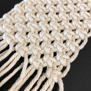 May include: A white macrame wall hanging with a fringe border. The macrame is made with a knotting technique that creates a grid pattern.