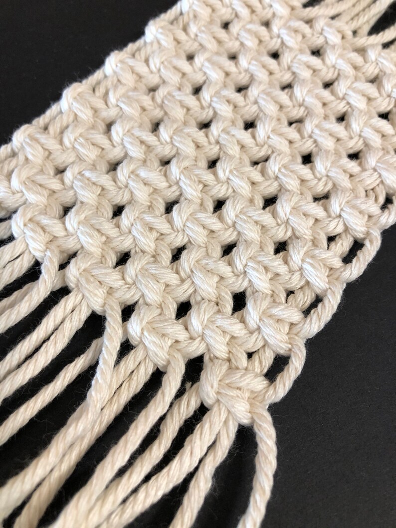 May include: A white macrame wall hanging with a fringe border. The macrame is made with a knotting technique that creates a grid pattern.