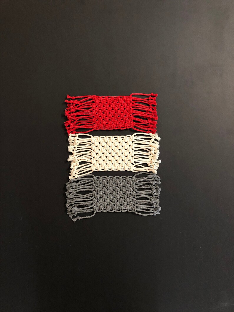 May include: Three handmade macrame coasters in red, white, and gray. The coasters are square and have a fringe border.