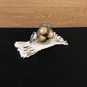 May include: A clear glass jar with a silver lid filled with gold-wrapped chocolate candies. The jar is sitting on a white crocheted coaster.