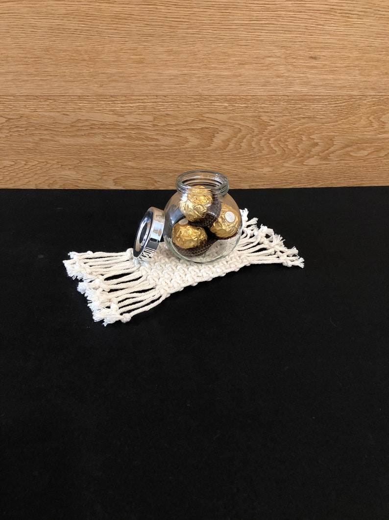 May include: A clear glass jar with a silver lid filled with gold-wrapped chocolate candies. The jar is sitting on a white crocheted coaster.
