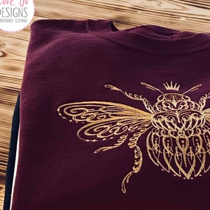 Puede incluir: Sudadera color burdeos con un diseño de abeja bordado en dorado. La abeja es intrincada y detallada, con una corona en la cabeza. La sudadera está doblada y apilada encima de otras dos sudaderas, una beige y otra negra.