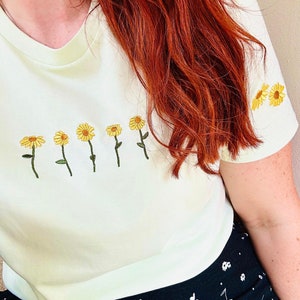 Peut inclure: T-shirt vert clair orn&eacute; de broderies de tournesols jaunes. Les fleurs sont dispos&eacute;es en rang&eacute;e sur le devant, avec d'autres sur la manche. Le t-shirt est port&eacute; avec une jupe noire &agrave; motifs floraux blancs.