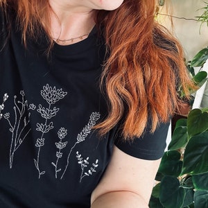 May include: Black t-shirt with a white embroidered floral design. The design features various flowers and stems, creating a botanical theme. The shirt has short sleeves and a crew neck.