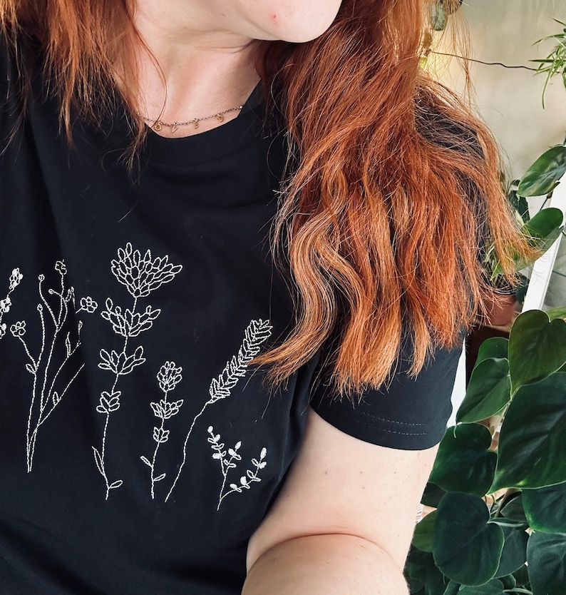 May include: Black t-shirt with a white embroidered floral design. The design features various flowers and stems, creating a botanical theme. The shirt has short sleeves and a crew neck.