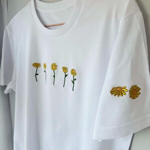 Peut inclure: T-shirt blanc avec broderie de fleurs de marguerite jaunes. Le motif pr&eacute;sente une rang&eacute;e de marguerites sur le devant et deux marguerites sur la manche. Le t-shirt a un col rond classique et des manches courtes.