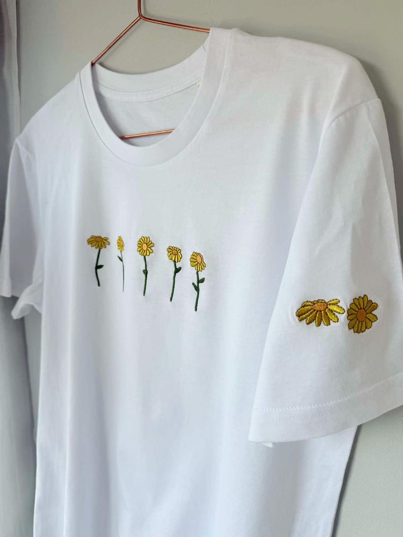 Peut inclure: T-shirt blanc avec broderie de fleurs de marguerite jaunes. Le motif pr&eacute;sente une rang&eacute;e de marguerites sur le devant et deux marguerites sur la manche. Le t-shirt a un col rond classique et des manches courtes.