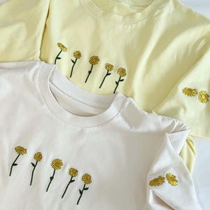Peut inclure: Deux t-shirts &agrave; manches courtes, un cr&egrave;me et un jaune p&acirc;le, sont pr&eacute;sent&eacute;s. Chaque chemise pr&eacute;sente une rang&eacute;e brod&eacute;e de marguerites jaunes avec des tiges vertes sur la poitrine. Le t-shirt jaune a une broderie de marguerite suppl&eacute;mentaire sur la manche.