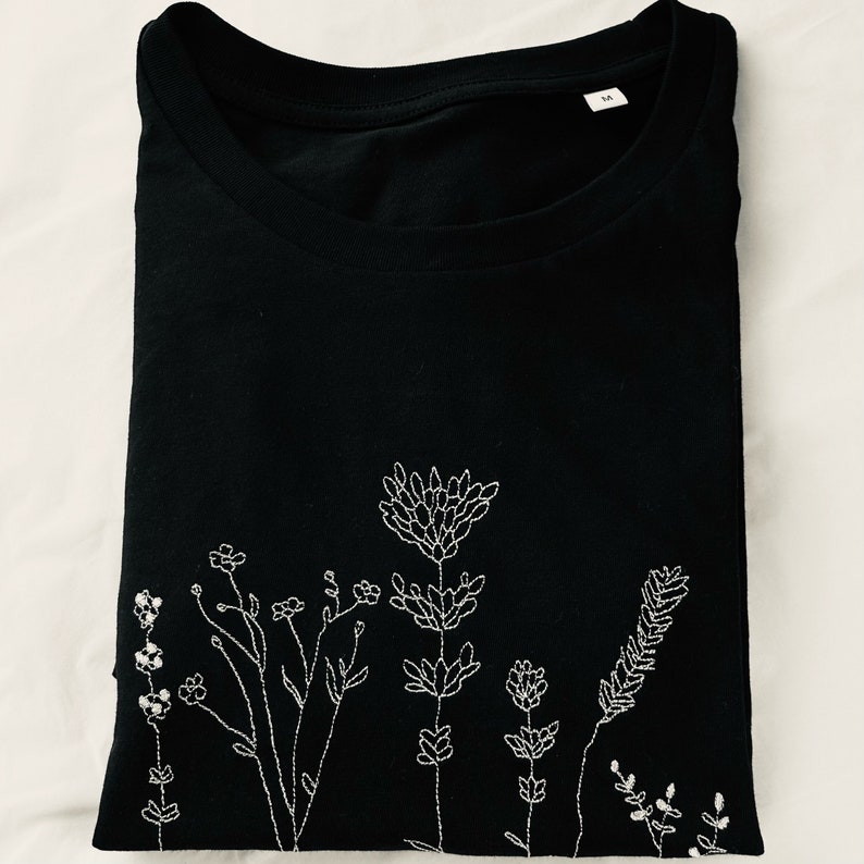 May include: Black t-shirt with a crew neck and short sleeves. The shirt has a white embroidered floral design across the lower front. The design features various stylized flowers and foliage.
