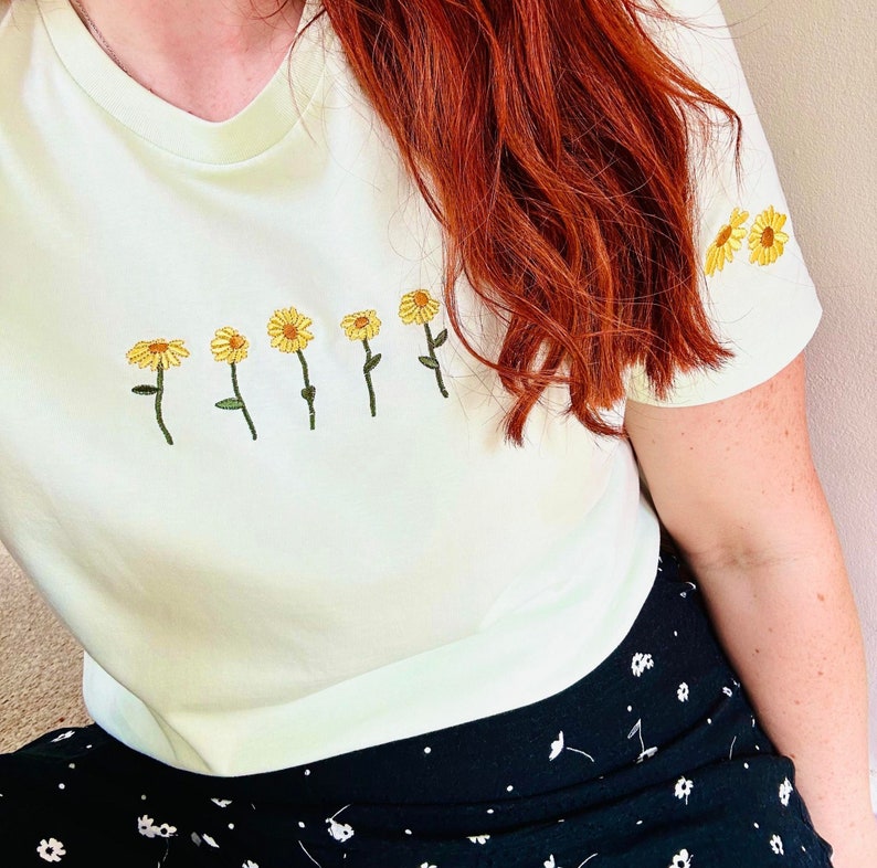 Peut inclure: T-shirt vert clair orn&eacute; de broderies de tournesols jaunes. Les fleurs sont dispos&eacute;es en rang&eacute;e sur le devant, avec d'autres sur la manche. Le t-shirt est port&eacute; avec une jupe noire &agrave; motifs floraux blancs.