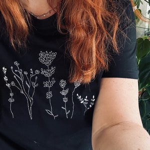 May include: Black t-shirt with a white embroidered floral pattern. The design showcases different stylized flowers and stems. The shirt has short sleeves and a round neck. The style is simple and elegant, suitable for everyday wear.