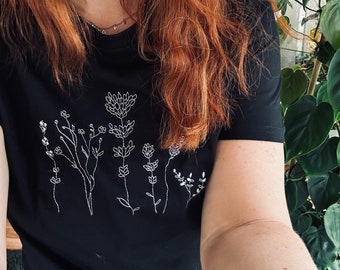 May include: Black t-shirt featuring a white embroidered floral design. The design includes various stylized flowers and stems. The shirt has short sleeves and a crew neck. The overall aesthetic is simple and elegant, suitable for casual wear.
