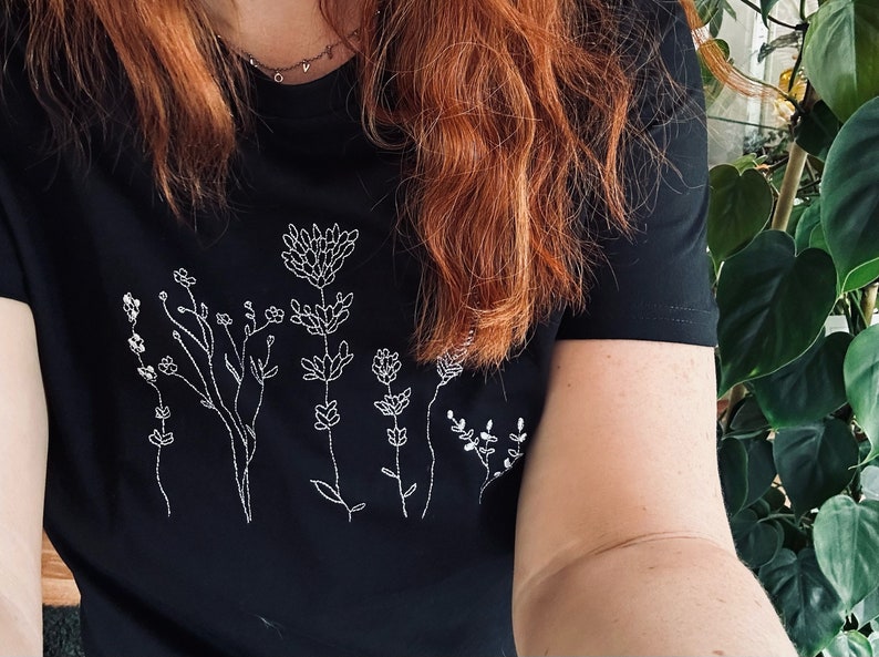 May include: Black t-shirt with a white embroidered floral pattern. The design showcases different stylized flowers and stems. The shirt has short sleeves and a round neck. The style is simple and elegant, suitable for everyday wear.