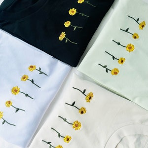 Peut inclure: Quatre t-shirts noirs, blancs, vert clair et cr&egrave;me. Chaque t-shirt pr&eacute;sente une broderie de tournesols jaunes avec des tiges vertes. Les t-shirts sont pli&eacute;s, mettant en valeur la broderie florale.