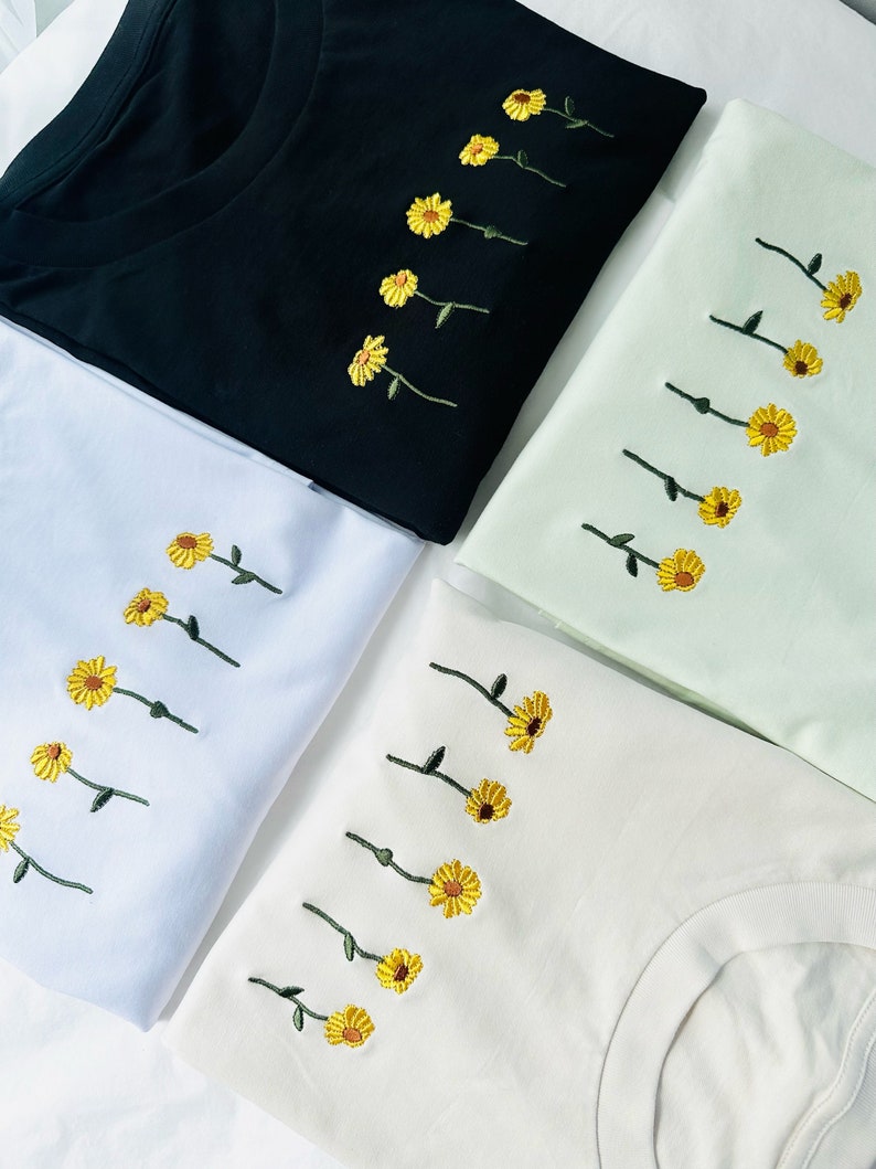 Peut inclure: Quatre t-shirts noirs, blancs, vert clair et cr&egrave;me. Chaque t-shirt pr&eacute;sente une broderie de tournesols jaunes avec des tiges vertes. Les t-shirts sont pli&eacute;s, mettant en valeur la broderie florale.
